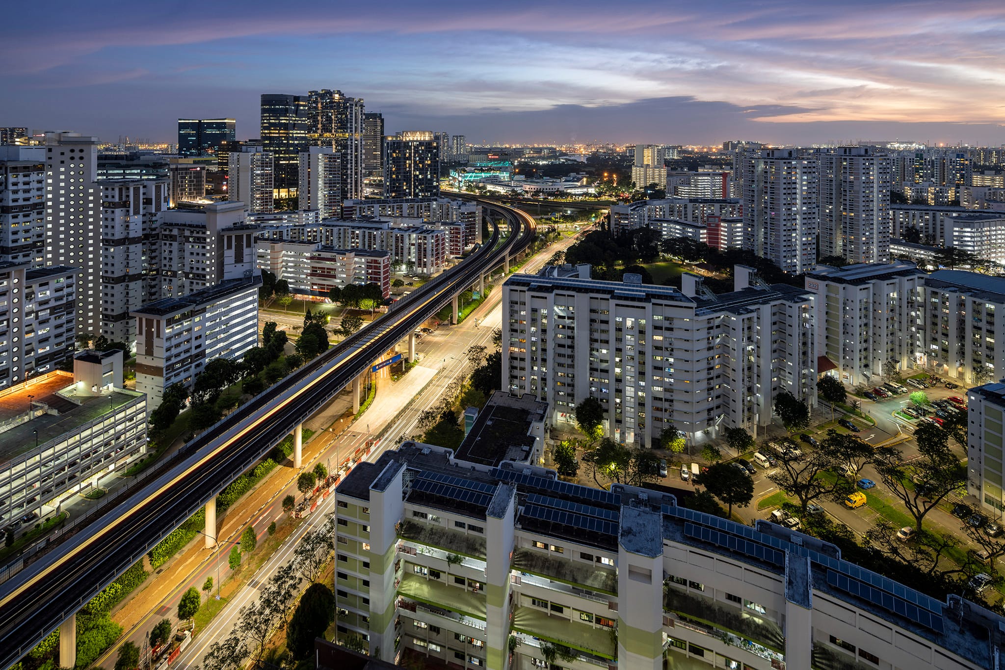 Jurong East at dusk with numerous high-rise buildings, an elevated train track, and roads illuminated by streetlights.